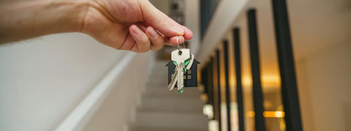 A hand holding a set of house keys on a house-shaped keychain in a bright entryway.